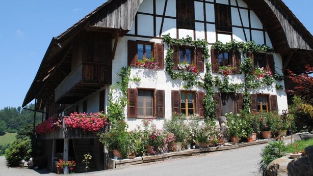 Ein traditionelles Fachwerkhäuschen mit einem steilen Dach, großen Fenstern mit Fensterläden und einem Balkon mit Blumenkörben.