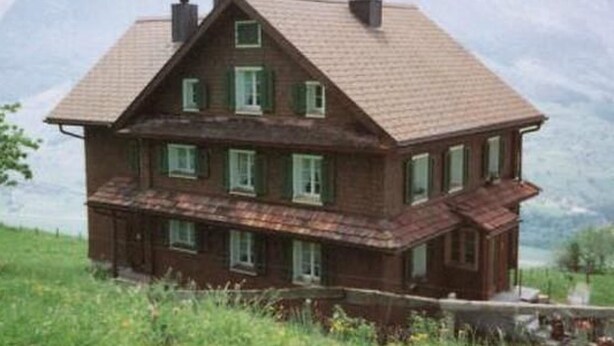 A two-story wooden house with a sloped roof, multiple windows, and a balcony.