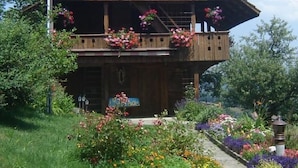 Eine Holzchalet mit Balkon, umgeben von einem Garten mit verschiedenen Blumen und Pflanzen.
