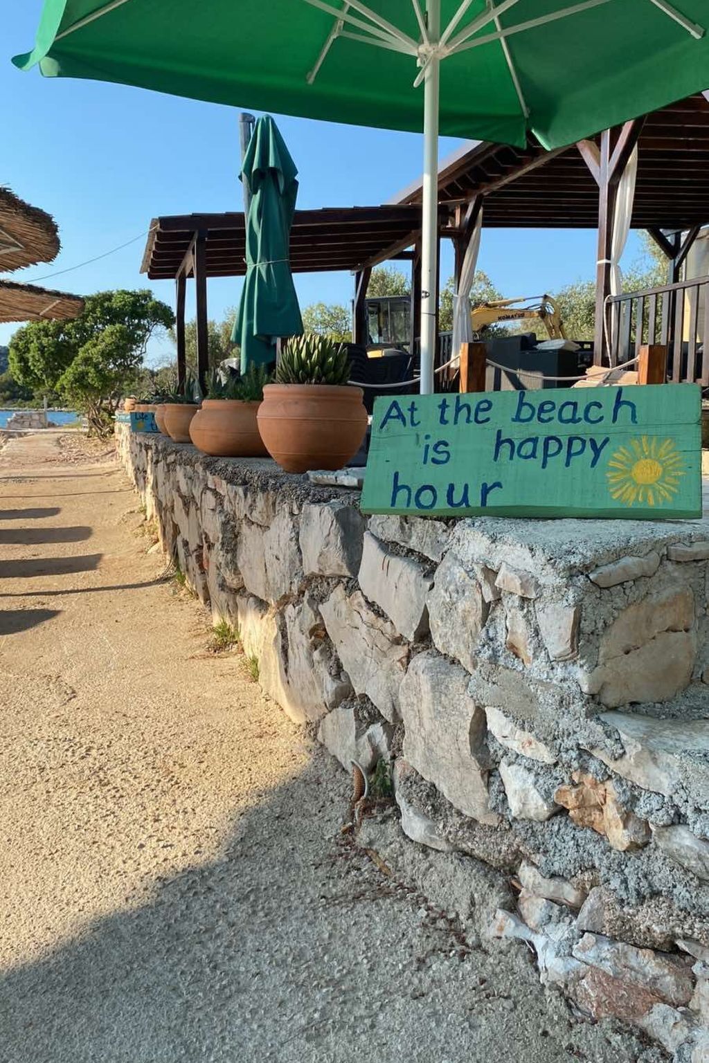 Un cadre idyllique en bord de mer, avec un mur de pierres, des plantes en pot et un panneau indiquant : « L’heure du bonheur, c’est à la plage. »
