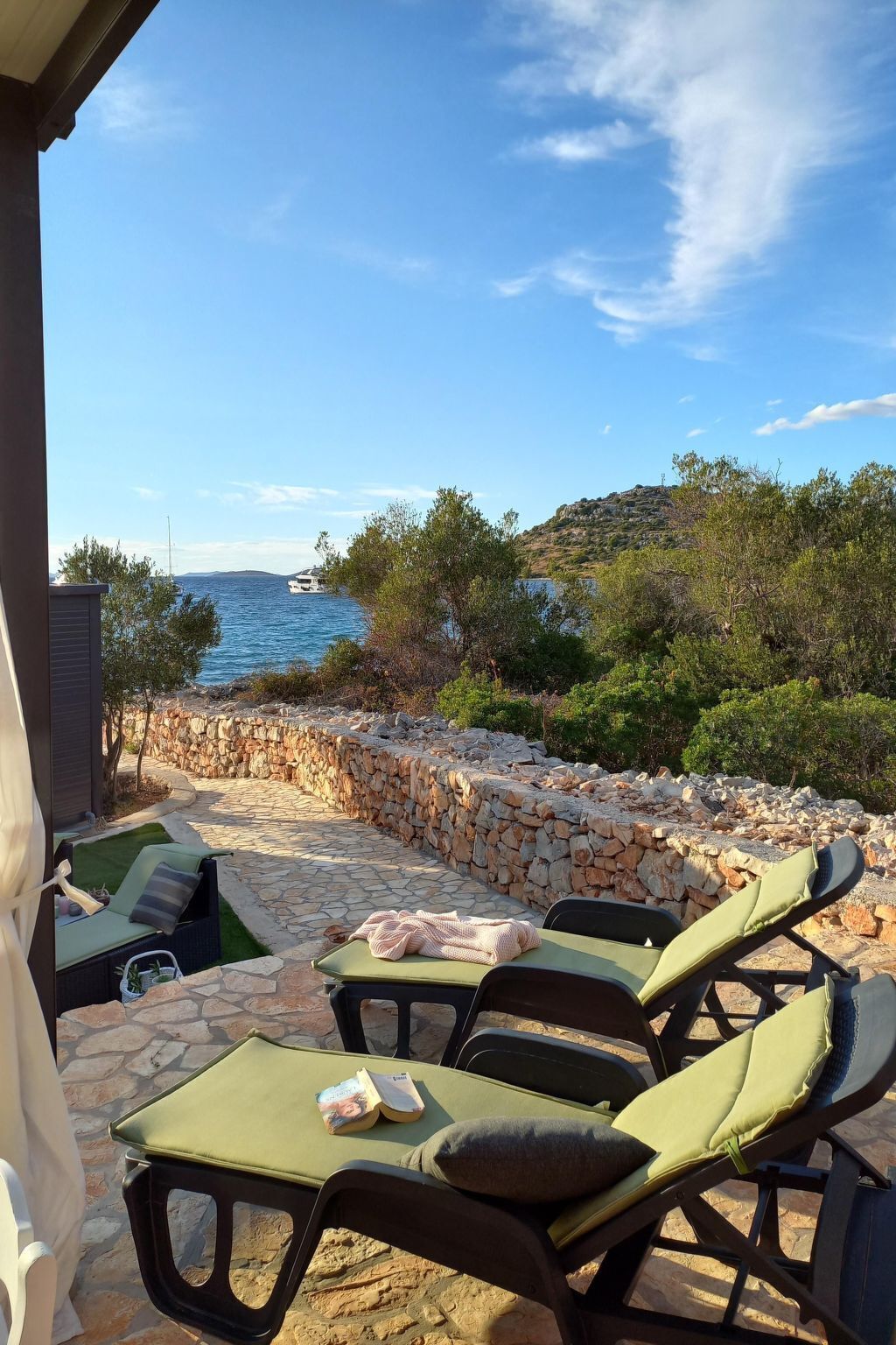 Une terrasse dotée de chaises longues et d’un mur en pierre, offrant une vue sur une étendue d’eau et un paysage vallonné.