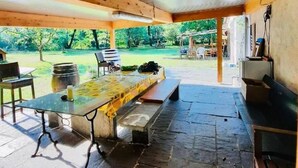 Un espace extérieur aménagé en terrasse, doté d’un sol en pierre, de poutres en bois au plafond et offrant une vue sur un jardin.