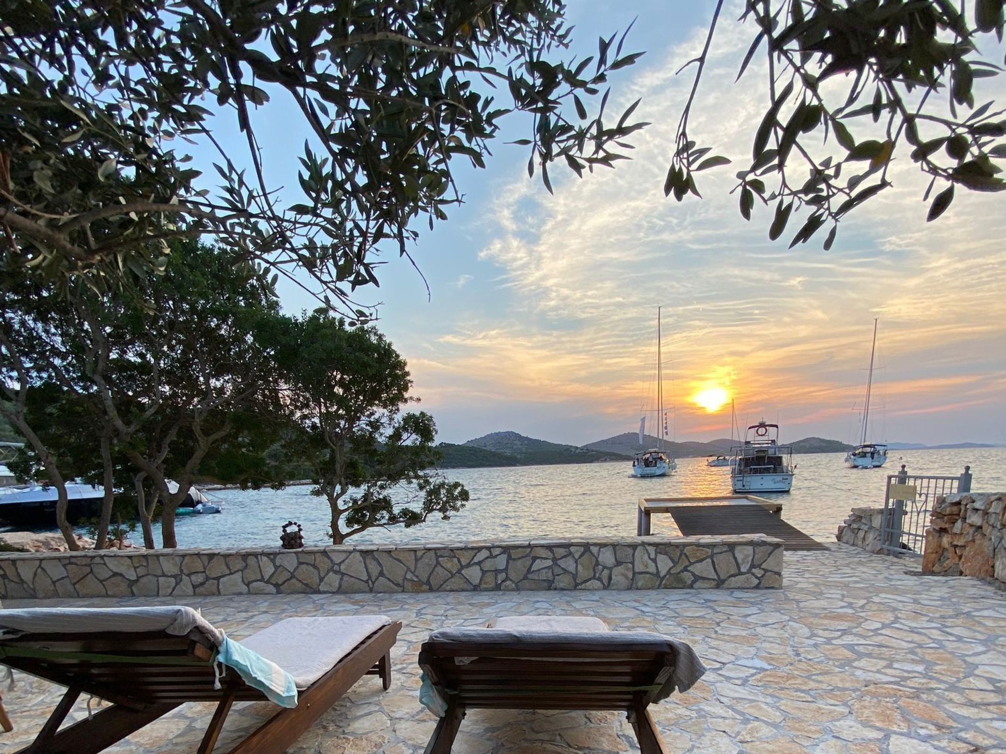 Un espace extérieur pavé de pierres, avec deux fauteuils de jardin en bois, un mur de pierres et une vue sur les bateaux sur l’eau au coucher du soleil.