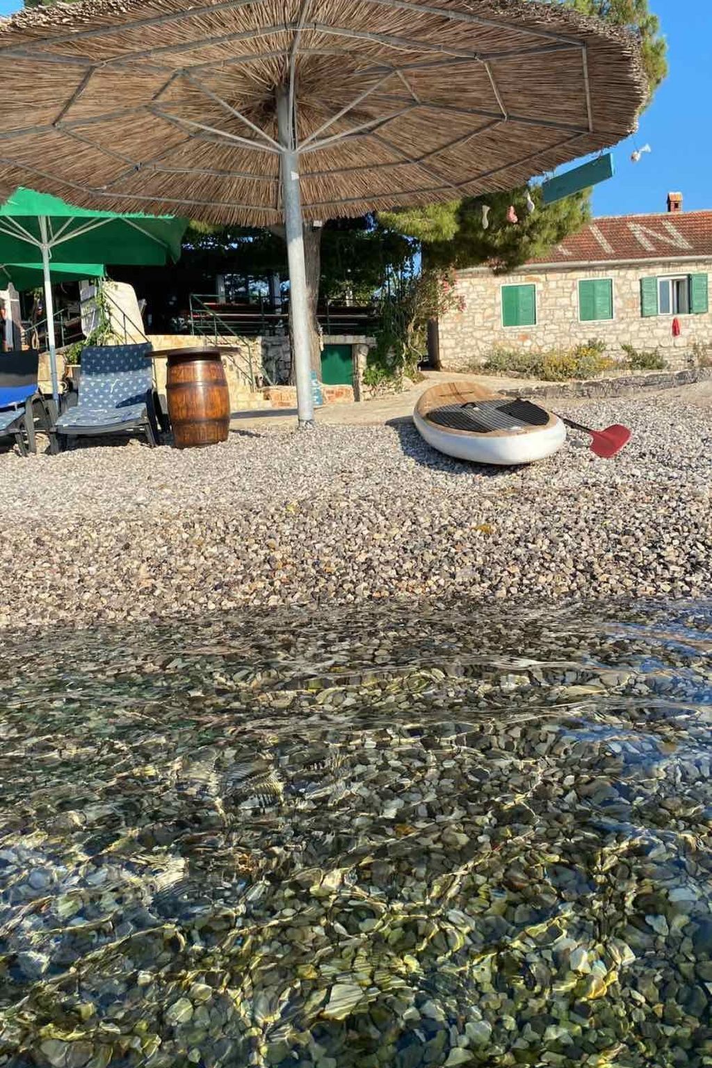 A pebbled beach area with a thatched umbrella, a paddleboard, and a stone building in the background.