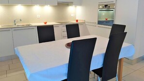 Une cuisine moderne avec des armoires blanches, un évier en acier inoxydable et un four encastré. On y trouve une table à manger avec quatre chaises.