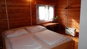 A wooden cabin room with a bed, a small table, a window with curtains, and a radiator. - Haus Cervená nad Vltavou mit Terrasse by Interhome (Jetetice)