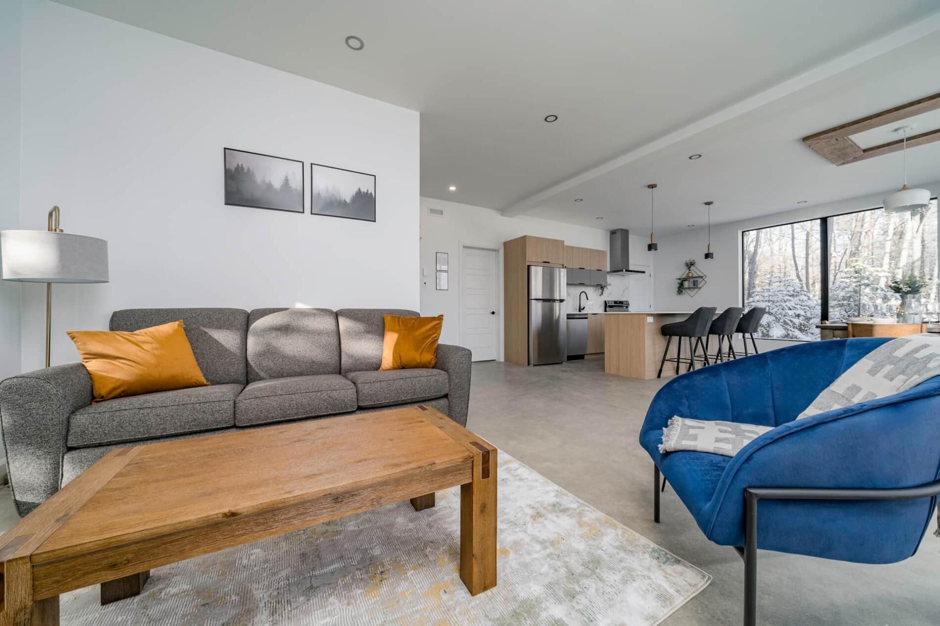 Een moderne woonkamer met een grijze bank, een houten salontafel en een blauwe fauteuil. Achtergrond toont een keuken en een eethoek.