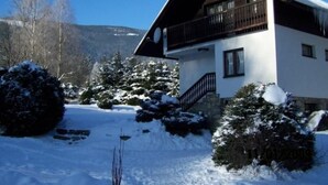 Un chalet de dos pisos con un balcón, rodeado de árboles cubiertos de nieve y una montaña al fondo.