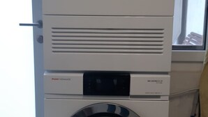 Un lave-linge à chargement frontal doté d’un panneau de commande numérique, situé dans une buanderie avec une fenêtre et une porte.