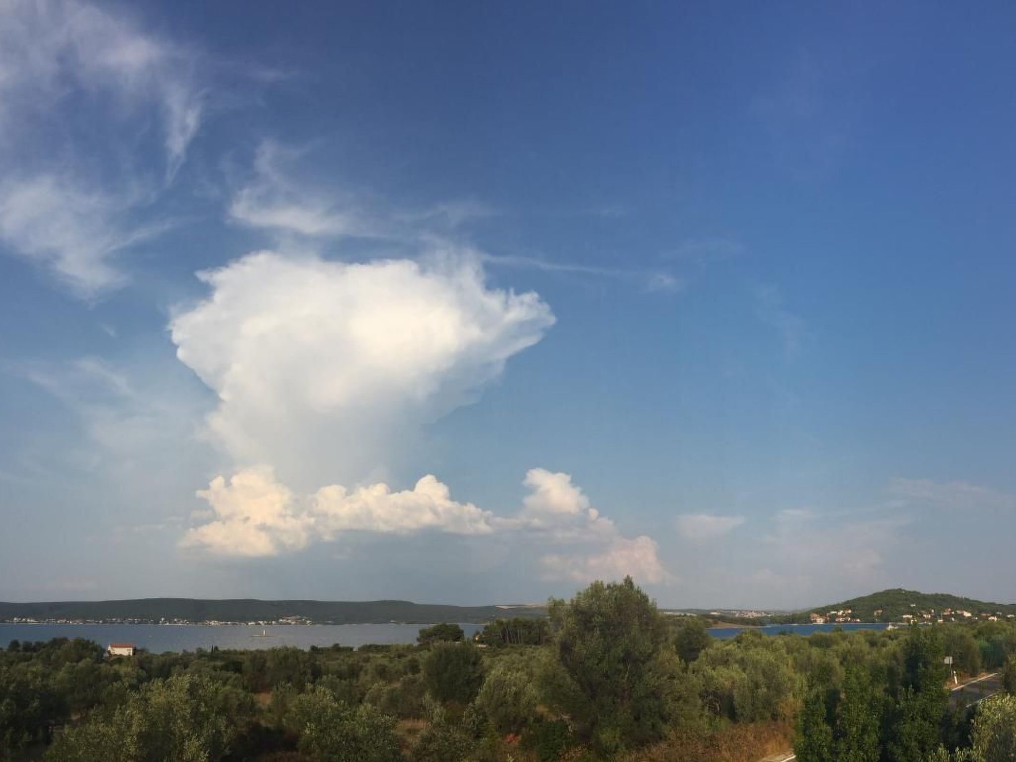 Paesaggio costiero con una vasta formazione nuvolosa, vegetazione lussureggiante e una vista lontana di una cittadina sull'acqua.