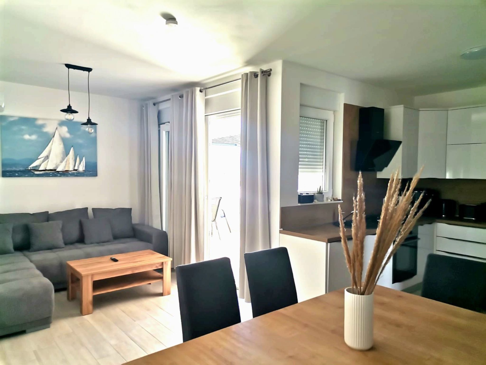 A modern living room with a gray sectional sofa, a wooden coffee table, and a dining table with chairs. There is a painting of sailboats on the wall, a ceiling light, and a kitchen area visible through an open door.