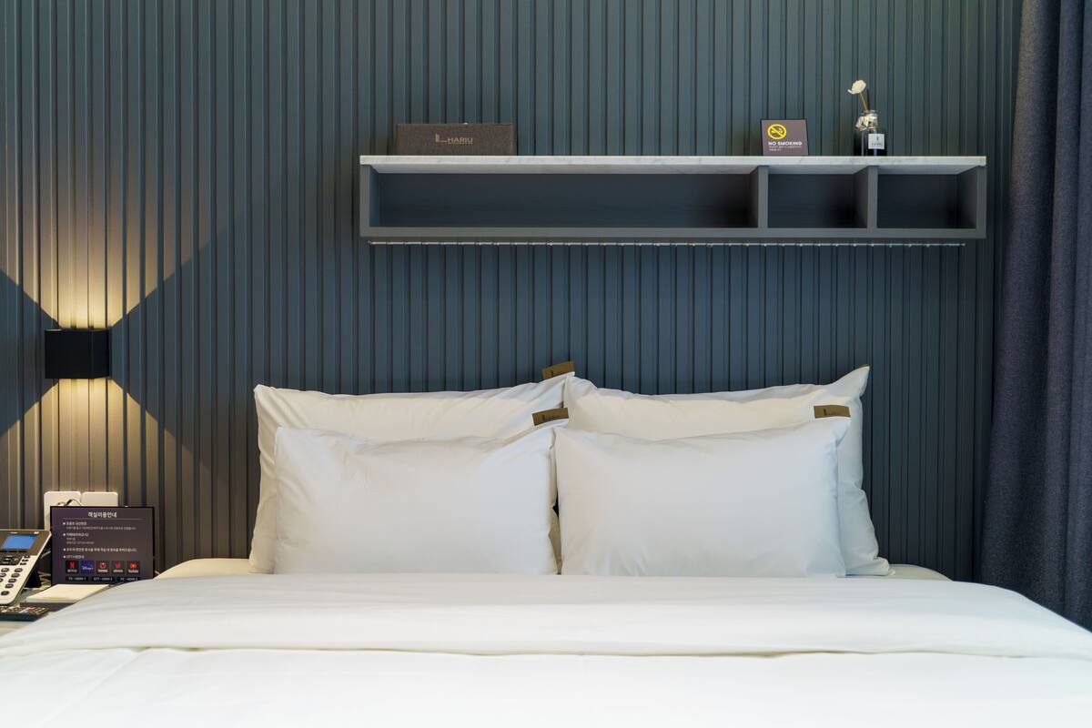 Habitación de hotel con cama, mesita de noche con teléfono, estante con un cartel y pared con líneas verticales.