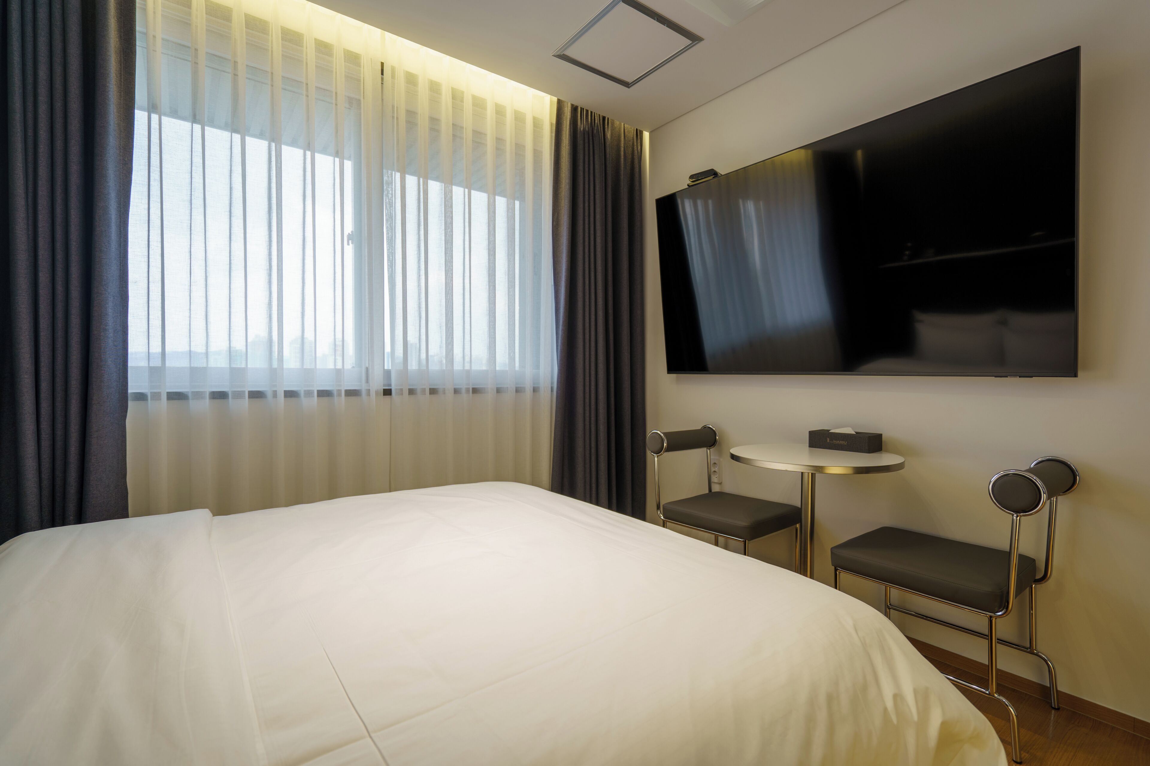 Hotellihuone, jossa on sänky, seinälle asennettu televisio, pieni pöytä ja kaksi tuolia.
