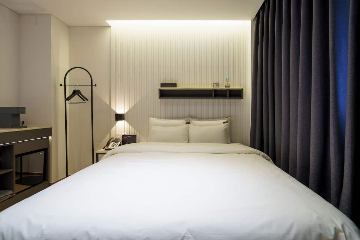 Habitación de hotel con cama, escritorio, teléfono, estante con un reloj y cortina.