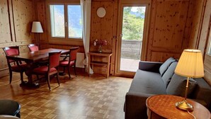 A wooden cabin interior with a dining table, chairs, a sofa, a lamp, and a view of the outdoors. - Casa Bambi 6.5 Zimmer by Interhome (Pfäfers)