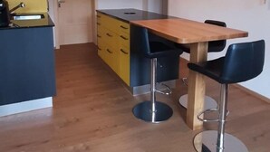 Une cuisine moderne avec des poutres en bois, un îlot central, des armoires jaunes et deux tabourets de bar noirs.