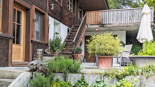 Une maison avec un escalier en bois, un balcon avec une chaise et un parasol, et un jardin avec différentes plantes.