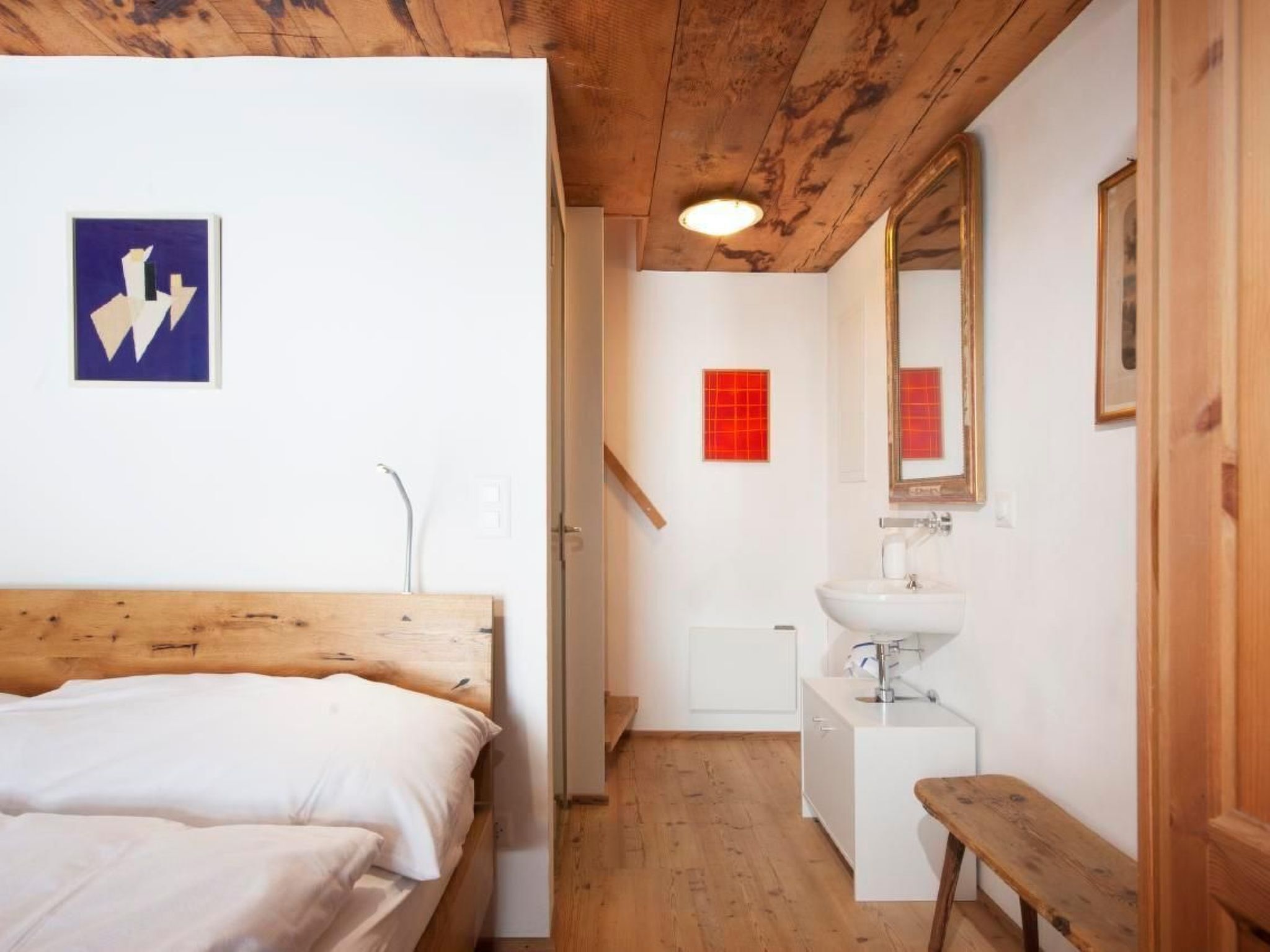 Un dormitorio con cabecera de madera, una cama, una mesita de noche, una pared blanca con un cuadro enmarcado, un adorno cuadrado rojo, un lavabo blanco, un espejo y un banco de madera.