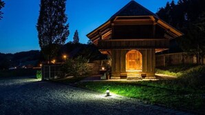 A small, wooden structure with a sloped roof, illuminated by warm lights, surrounded by a gravel path and greenery.