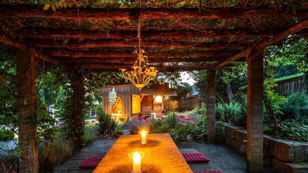 A covered outdoor area with a pathway leading to a building, surrounded by greenery and a warm light source.
