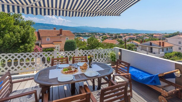 Une terrasse avec une table ronde dressée pour le dîner, d’où l’on peut admirer un quartier résidentiel avec ses maisons et, au loin, des montagnes.