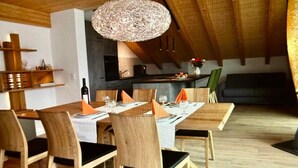 Comedor con techo alto y vigas de madera, con una mesa redonda preparada para cuatro personas, sillas de mimbre y una lámpara colgante.