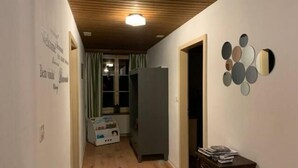 Un couloir étroit avec un plancher en bois, un luminaire au plafond, un réfrigérateur et une petite table sur laquelle sont posés des livres.