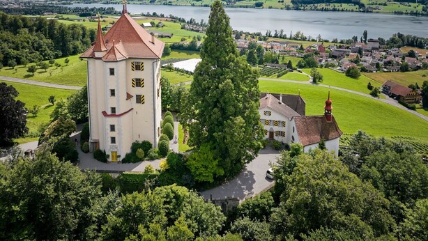 Un château coiffé d’un toit rouge, entouré de prairies verdoyantes et bordé d’un lac.