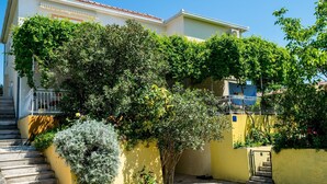 Un edificio residencial con una escalera que lleva a un balcón, rodeado de exuberante follaje y un muro amarillo.