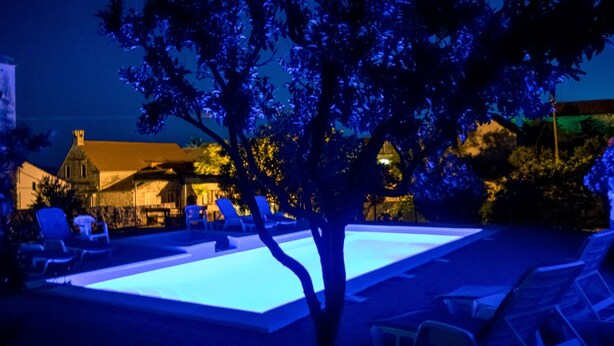 Escena nocturna con una piscina iluminada con luces azules, rodeada de sillas de descanso y un árbol.