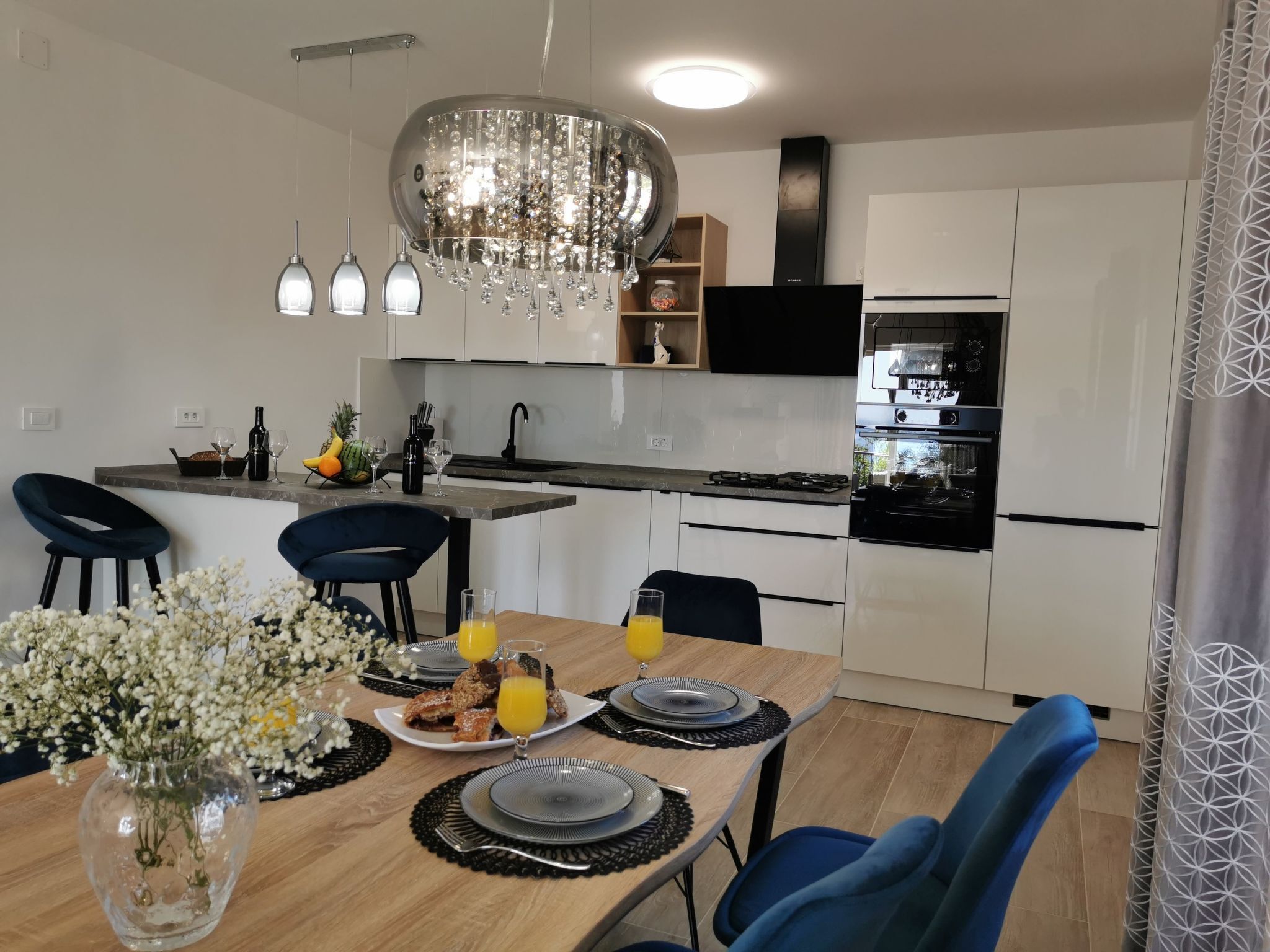 Une cuisine moderne avec des armoires blanches, une cuisinière noire et un four encastré. On y trouve également un coin repas avec une table en bois, des chaises bleues et un vase rempli de fleurs.
