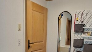 Une pièce avec une porte en bois, une armoire blanche avec des tiroirs, un miroir et un panneau perforé où sont accrochés des sacs.