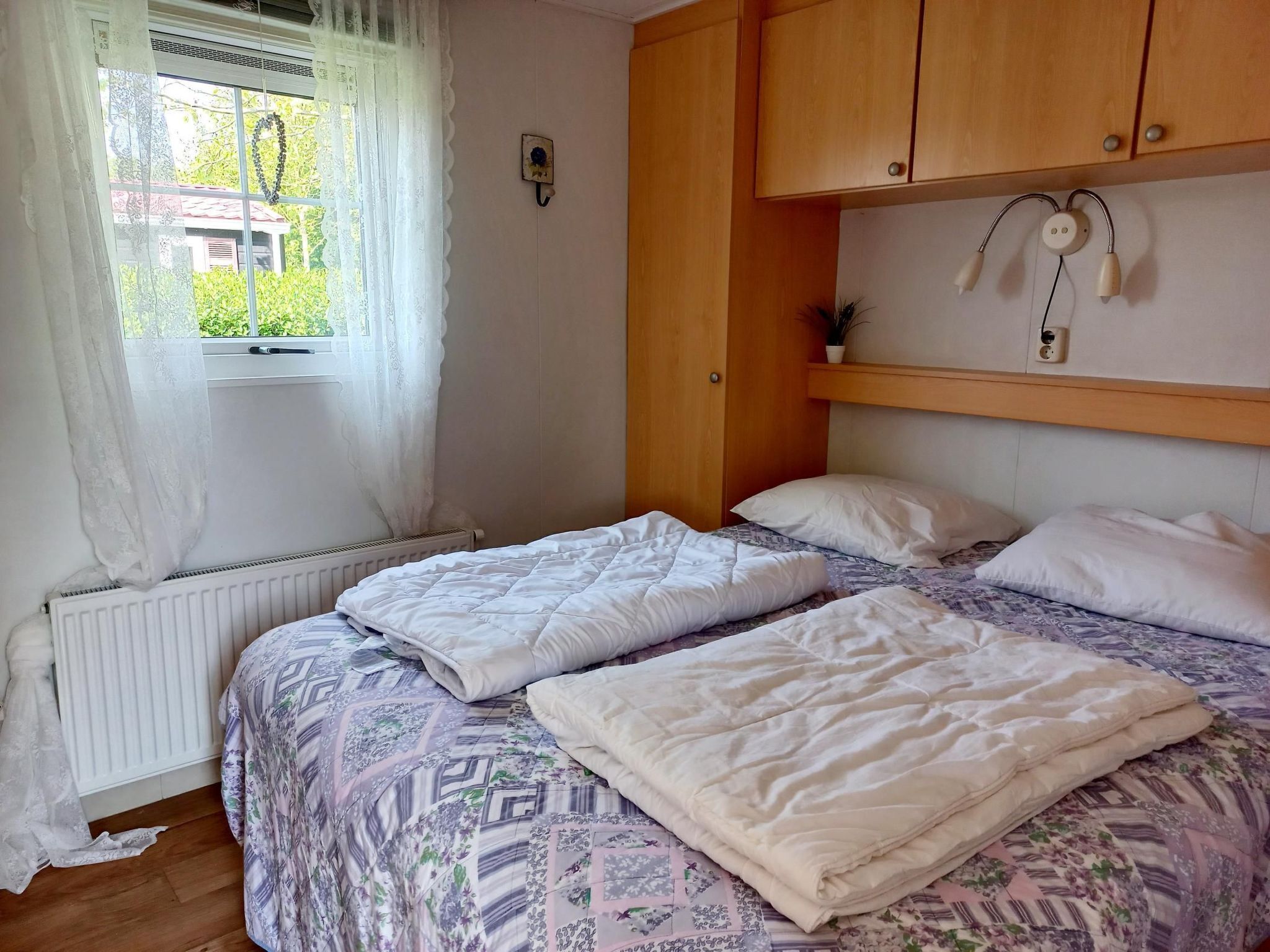Un dormitorio con dos camas, una ventana con cortinas de gasa, un radiador y armarios de madera sobre las camas.
