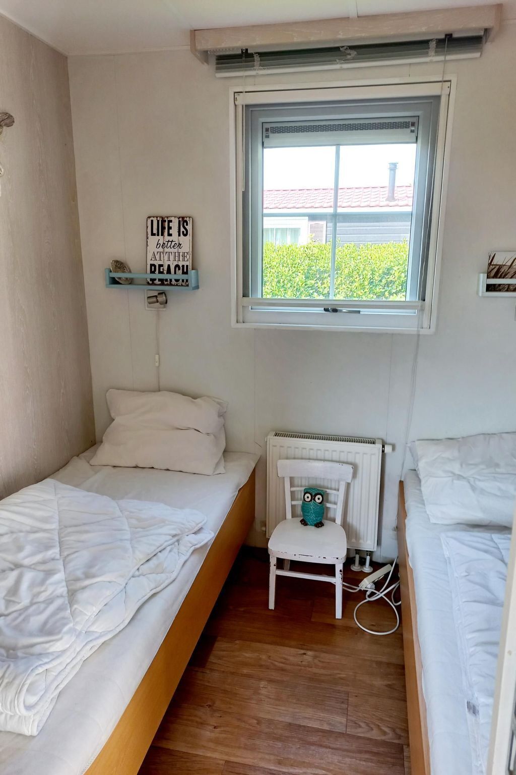 A small, cozy room with two single beds, a radiator, a window with a view of greenery, and a wall-mounted shelf with a decorative sign.