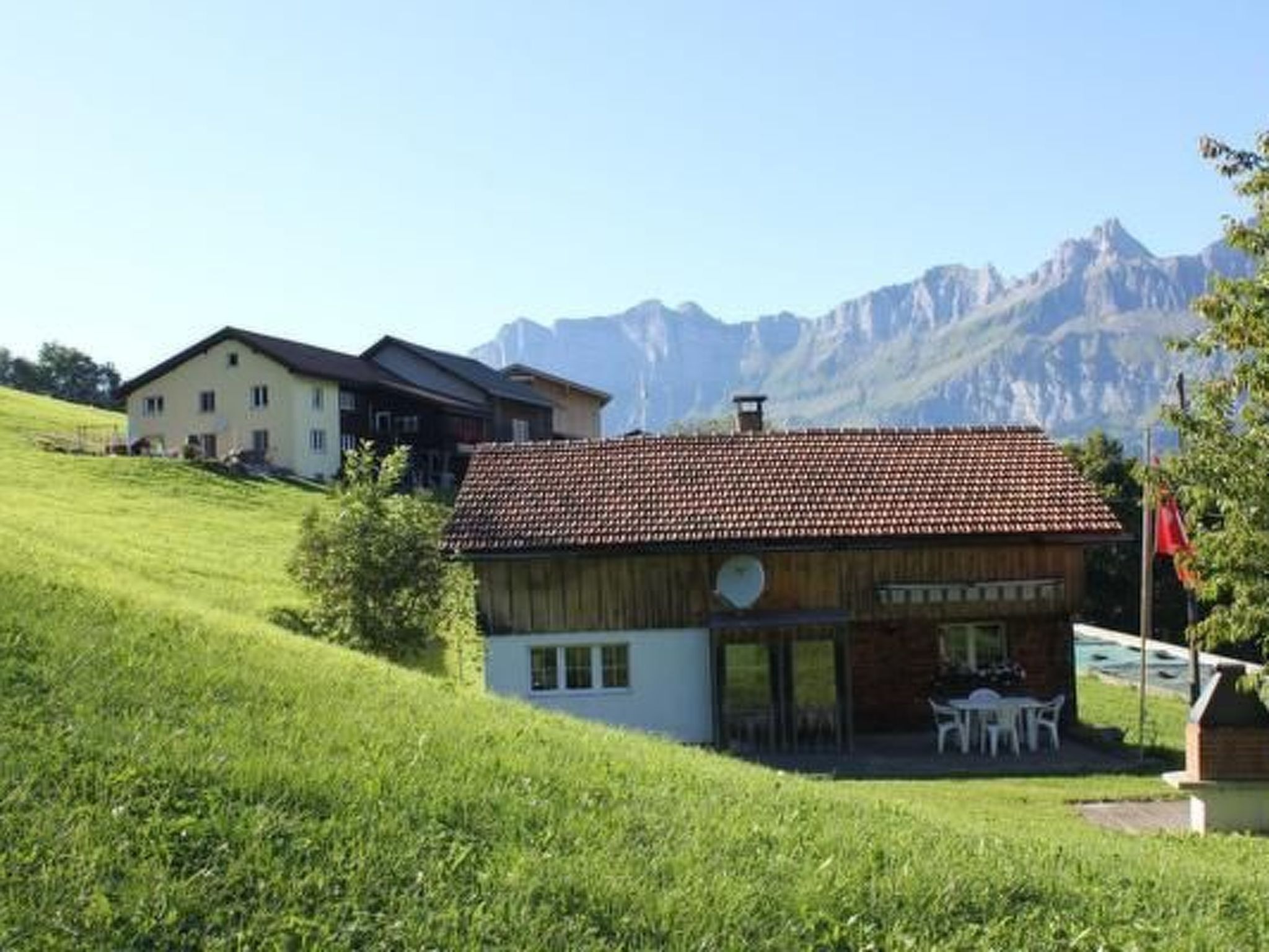 Una pequeña casa de madera con techo de tejas, un patio con mobiliario exterior y montañas al fondo.