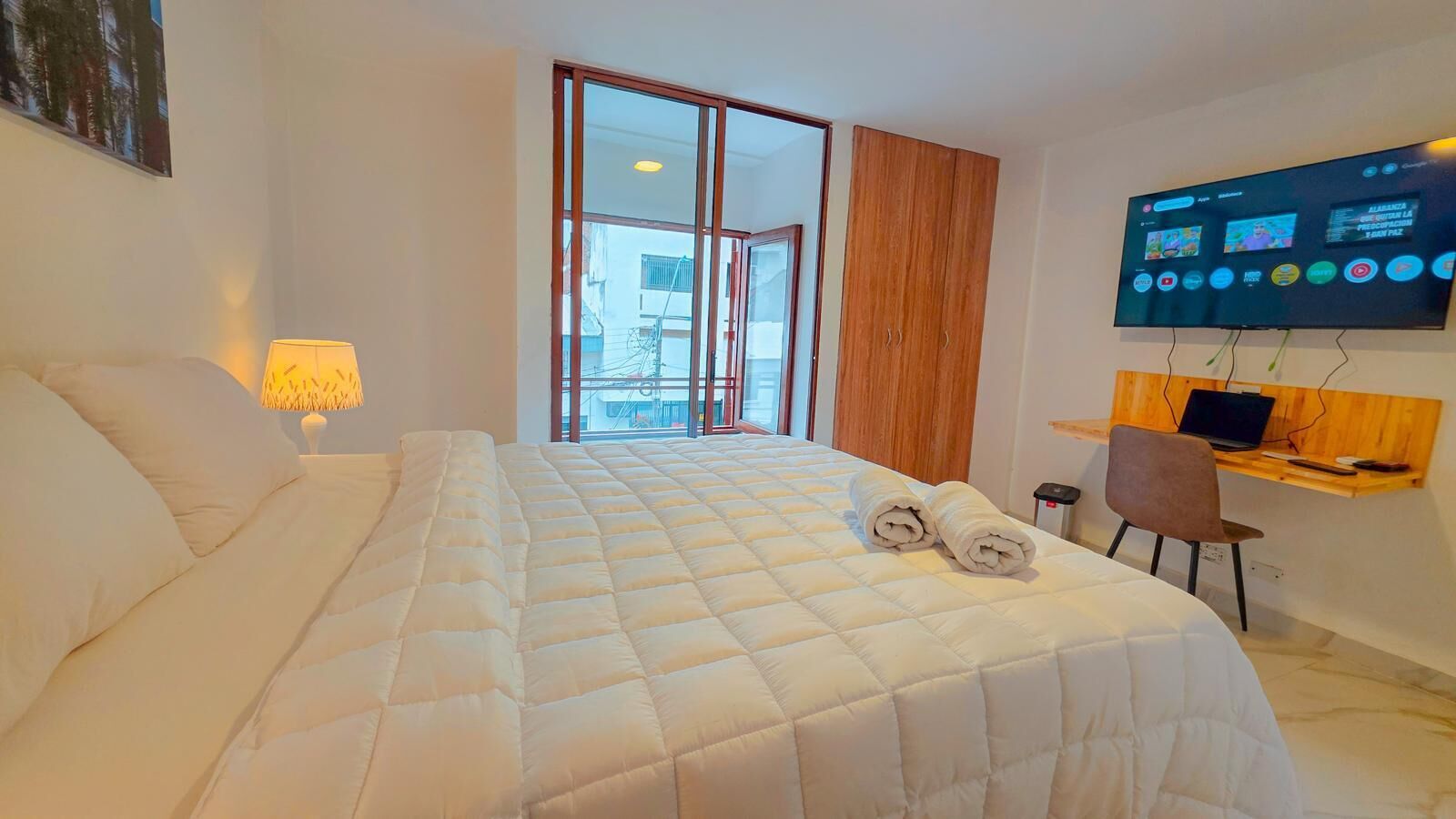 A hotel room with a large bed, a desk with a computer, and a TV on the wall.