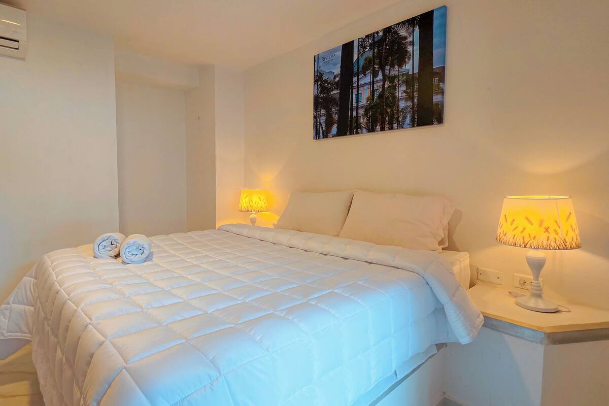 A hotel room with a white bed, two rolled towels, a painting on the wall, and two bedside lamps.