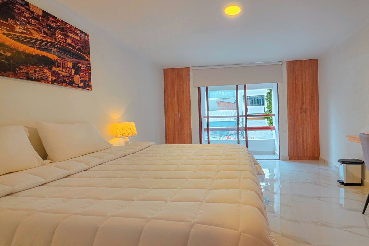 A hotel room with a bed, a desk with a computer, a chair, a lamp, and a large window.