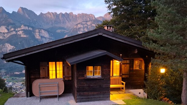 A wooden cabin with a porch, a bench, and a view of mountains.