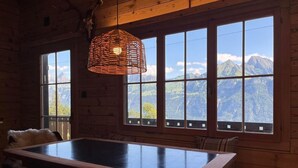 A wooden cabin interior with a large dining table, multiple windows offering mountain views, and a woven light fixture.