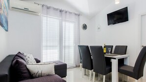 Ein modernes Wohnzimmer mit einer Couch, einem Esstisch mit Stühlen, einem an der Wand montierten Fernseher und einem fenster mit Vorhängen.