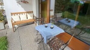 Un balcon avec un banc en bois, une table avec deux tasses, et une vue sur une voiture verte et des arbres.