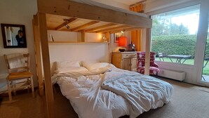 Une chambre avec un plafond en bois, un lit recouvert de linge de lit blanc, une chaise, un bureau avec une lampe et une vue sur l’extérieur par le biais d’une grande fenêtre.