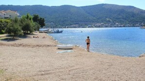 En strand med en person som står vid vattnet, båtar i fjärran och ett kuperat landskap i bakgrunden.