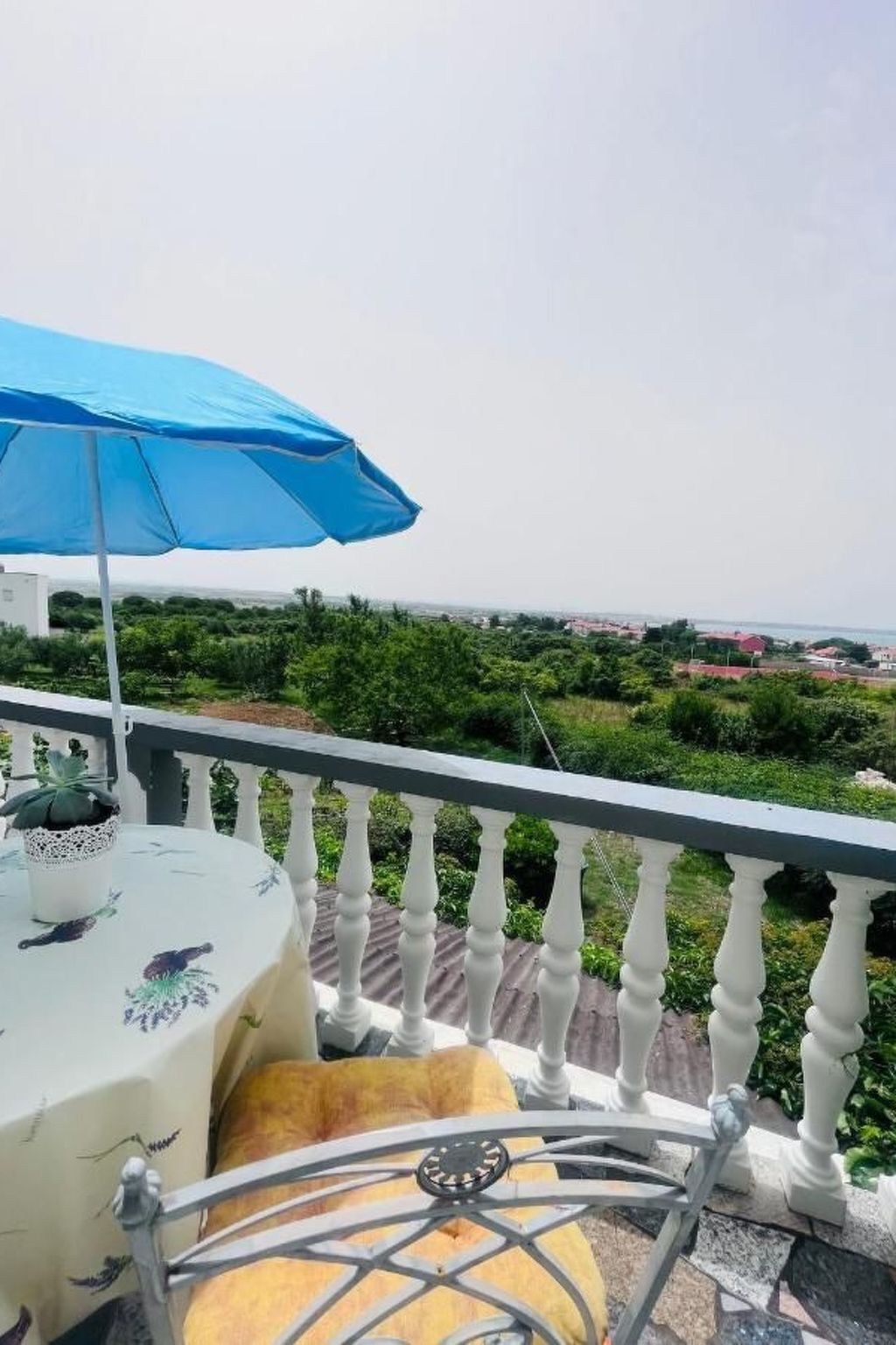 Ein Balkon mit einem blauen Sonnenschirm, einem Tisch mit einer blumigen Tischdecke und einem gelben Kissen.