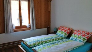 Een enkel bed met een kleurrijke deken en kussens met een geometrisch patroon, een houten vloer, een raam met gordijnen en een klein tafeltje met een lamp.