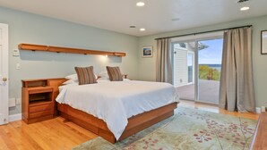 Un dormitorio con una cama grande, mesitas de noche de madera, una pequeña mesa y vistas por un ventanal corredero.