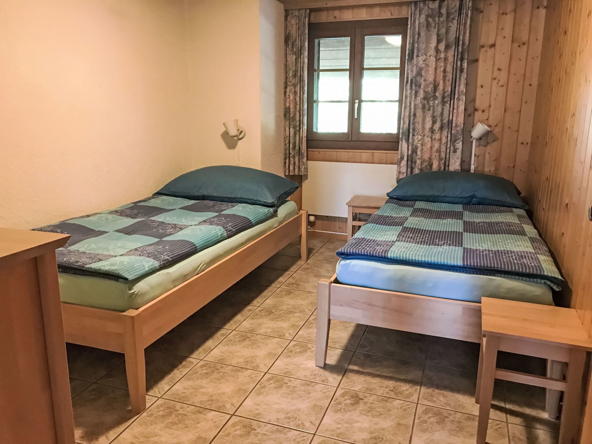 A room with two wooden beds, a window with curtains, a small table, and a wall-mounted light fixture.