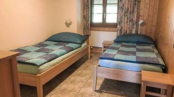 A room with two wooden beds, a window with curtains, a small table, and a wall-mounted light fixture.