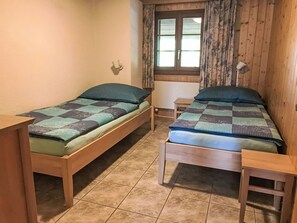 Ein Zimmer mit zwei Holzbett, einem Fenster mit Vorhängen, einem kleinen Tisch und einer Wandleuchte.